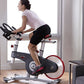 Woman in white tank top and black shorts riding black and red stationary exercise bike in bright home gym space