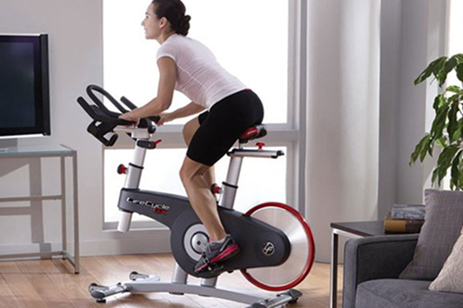 Woman in white tank top and black shorts riding black and red stationary exercise bike in bright home gym space