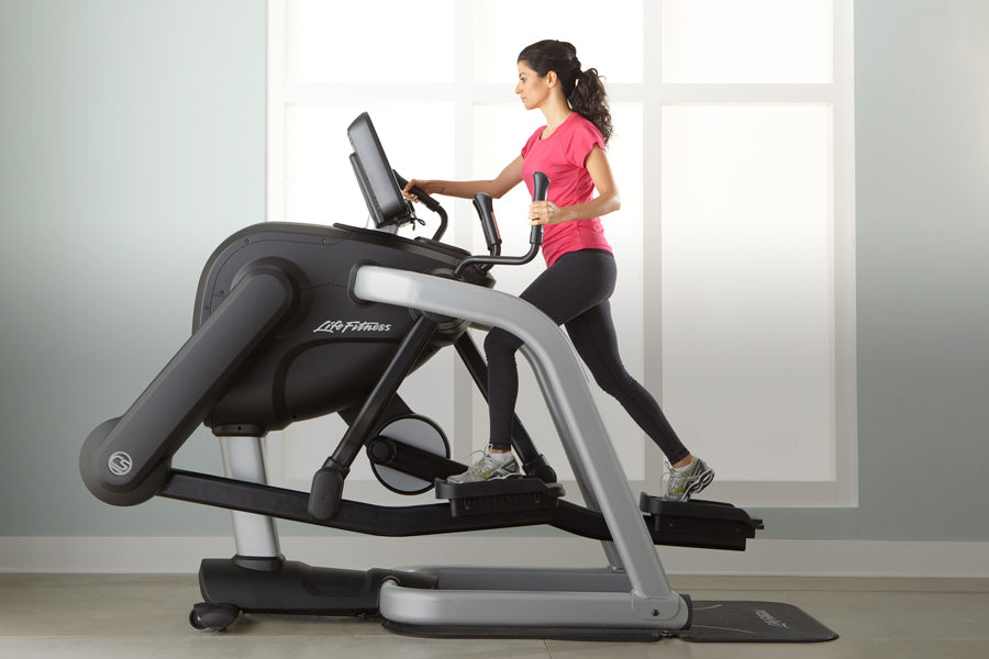 Woman in pink top and black leggings using Life Fitness elliptical trainer with digital display in bright studio setting