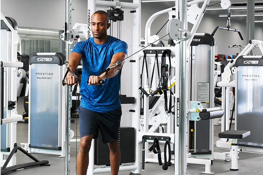 Man in blue t-shirt using Cybex Prestige dual adjustable pulley machine in gym setting surrounded by fitness equipment