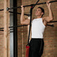 Man in white tank top using black pull-up bar with red resistance bands mounted on brick wall