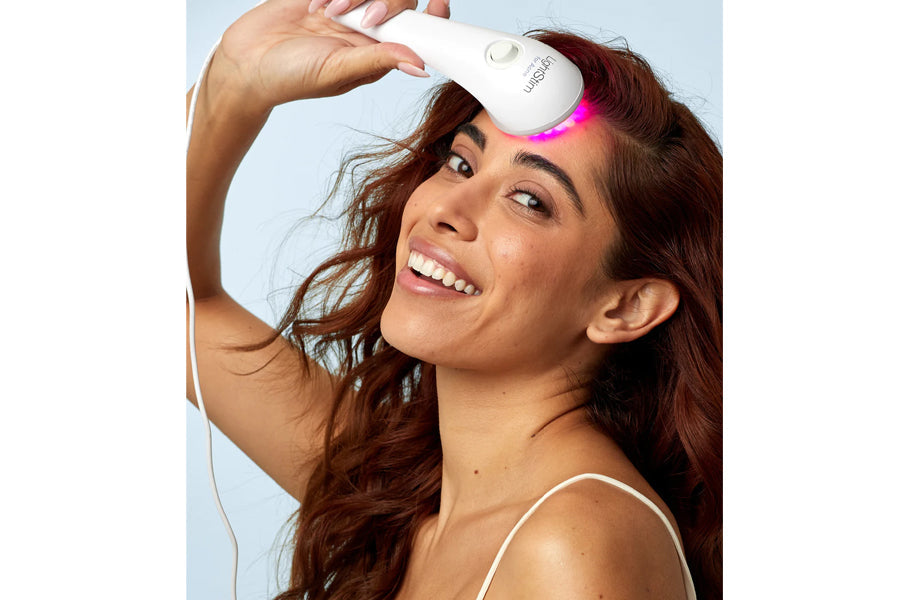 Woman with long reddish-brown hair holding white handheld LED light therapy device with pink light on forehead against blue b