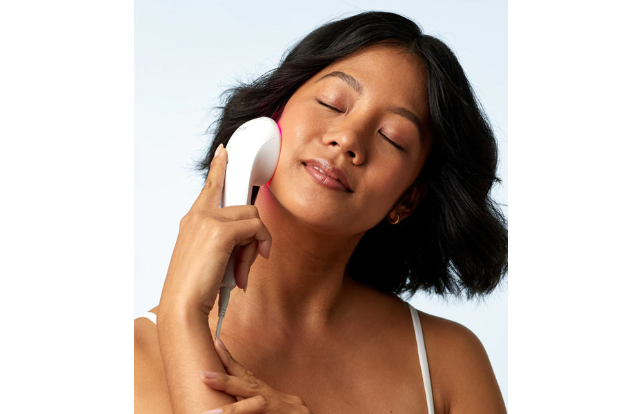 White handheld LED light therapy device used on cheek, worn by model with dark hair against light blue background