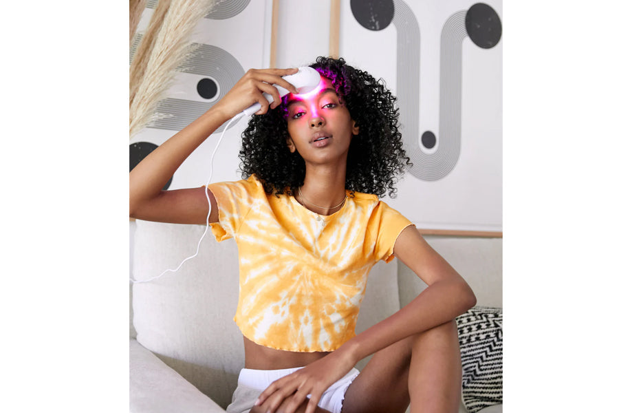 Model wearing yellow and white tie-dye crop t-shirt, holding pink LED light therapy device near face