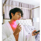 Person in white waffle-knit robe holding handheld LED light therapy device emitting pink light on neck and shoulder area in b