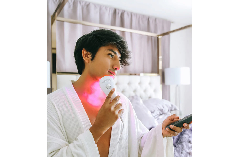 Person in white waffle-knit robe holding handheld LED light therapy device emitting pink light on neck and shoulder area in b