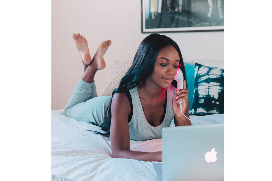 Woman lying on bed in gray sleeveless tank top using LightStim LED light therapy device while working on laptop
