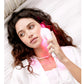White LED light therapy device with pink glow, held against model's cheek wearing white blazer and pearl necklace