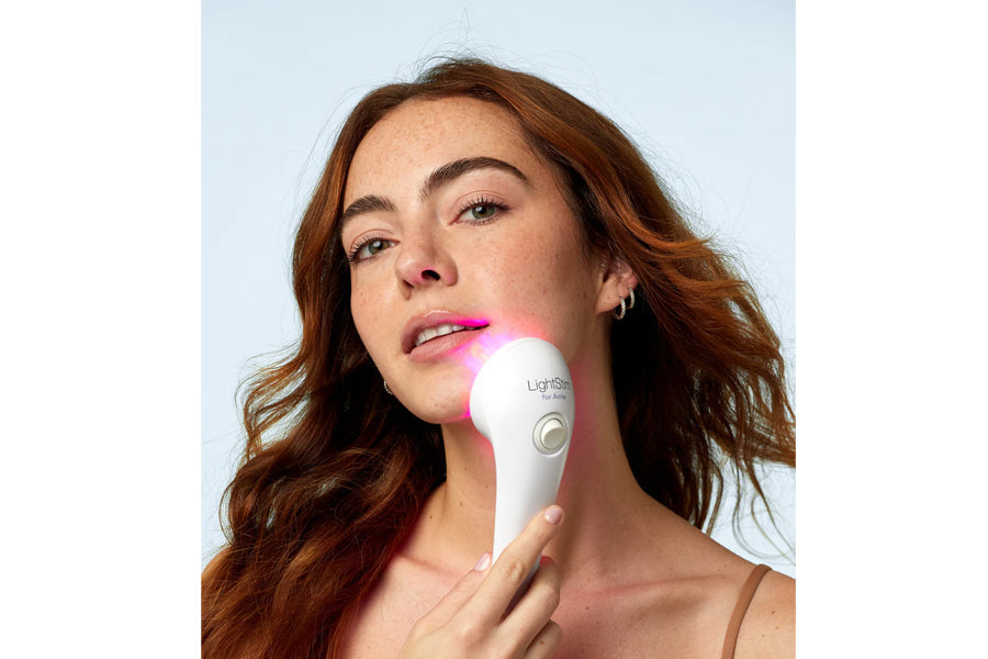 Woman with reddish-brown hair holding white LightStim LED light therapy device with pink light against chin, on light blue ba