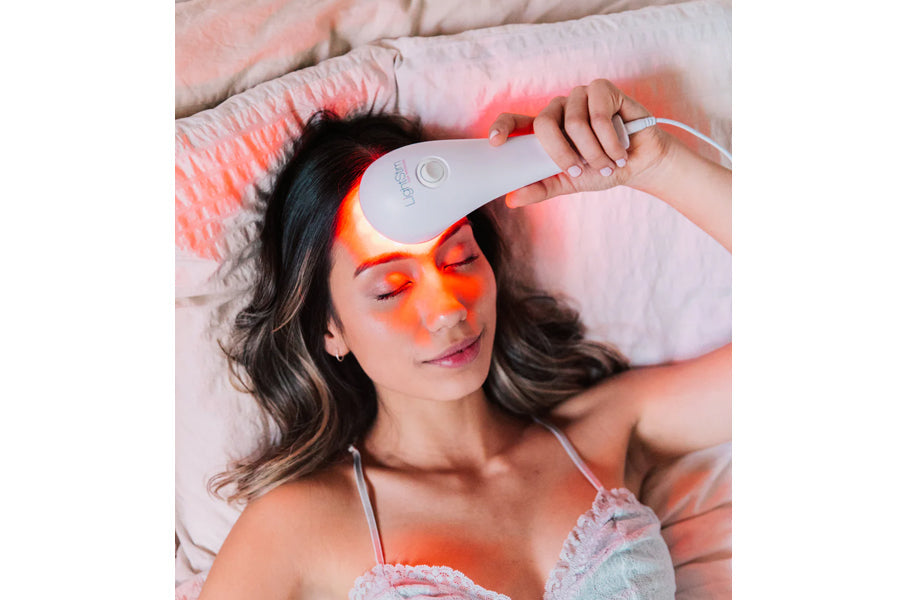 Woman using white LED light therapy device on forehead, emitting red and orange light, worn on bed with pink pillows