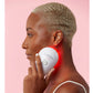 White handheld LED light therapy device with red light emitting from center, held against jawline by model on pink background