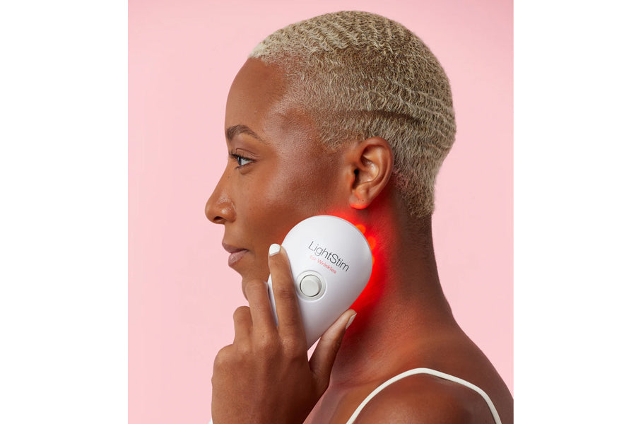 White handheld LED light therapy device with red light emitting from center, held against jawline by model on pink background