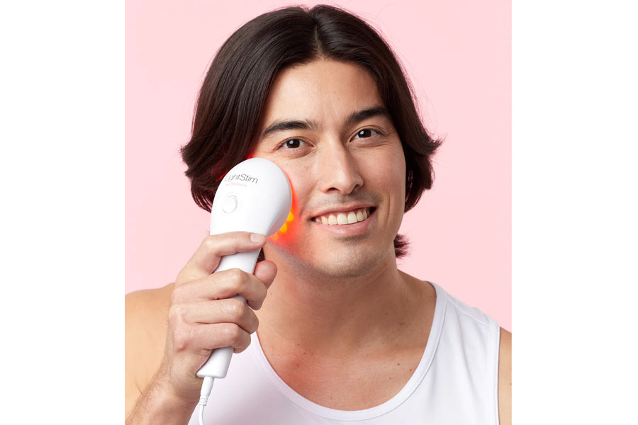 Man holding white LED light therapy device with red light glow near his face against pink background