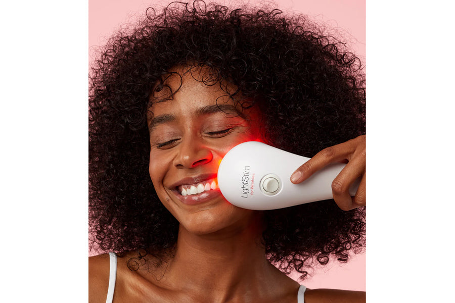 LightStim LED light therapy device with red light emitting from white handheld applicator, held against model's cheek on pink