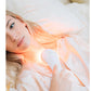 Woman with blonde hair holding white handheld LED light therapy device on chest, lying on white bedding with warm orange ligh