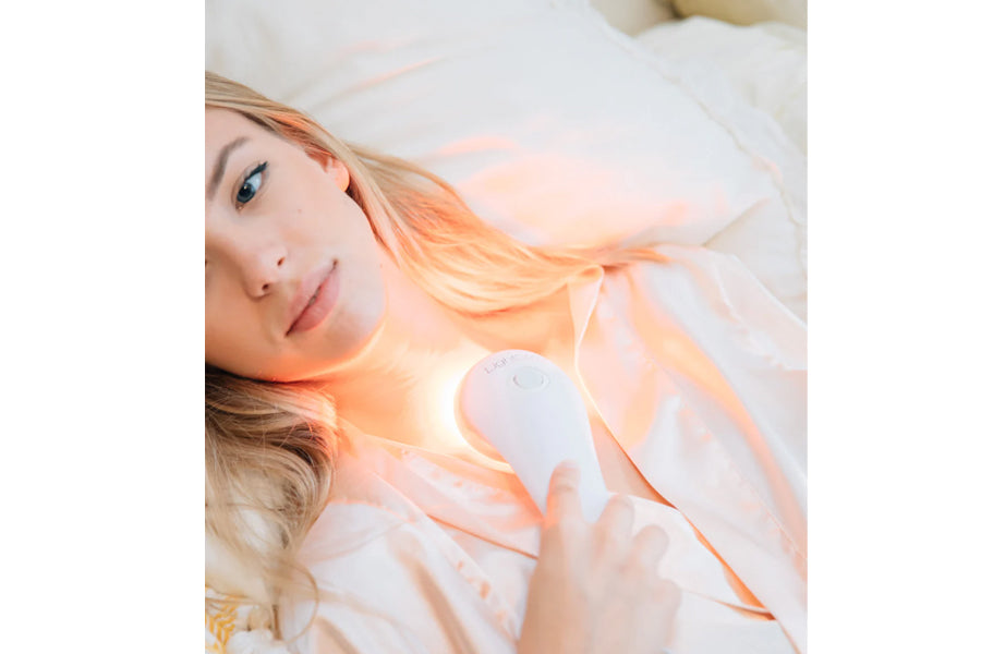 Woman with blonde hair holding white handheld LED light therapy device on chest, lying on white bedding with warm orange ligh