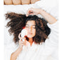 Woman with curly brown hair using white LED light therapy device on face, lying on white bedding