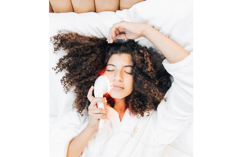 Woman with curly brown hair using white LED light therapy device on face, lying on white bedding