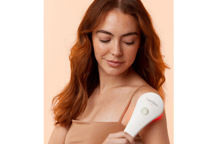Woman with red hair holding white and red LightStim LED therapy device against shoulder on peach background