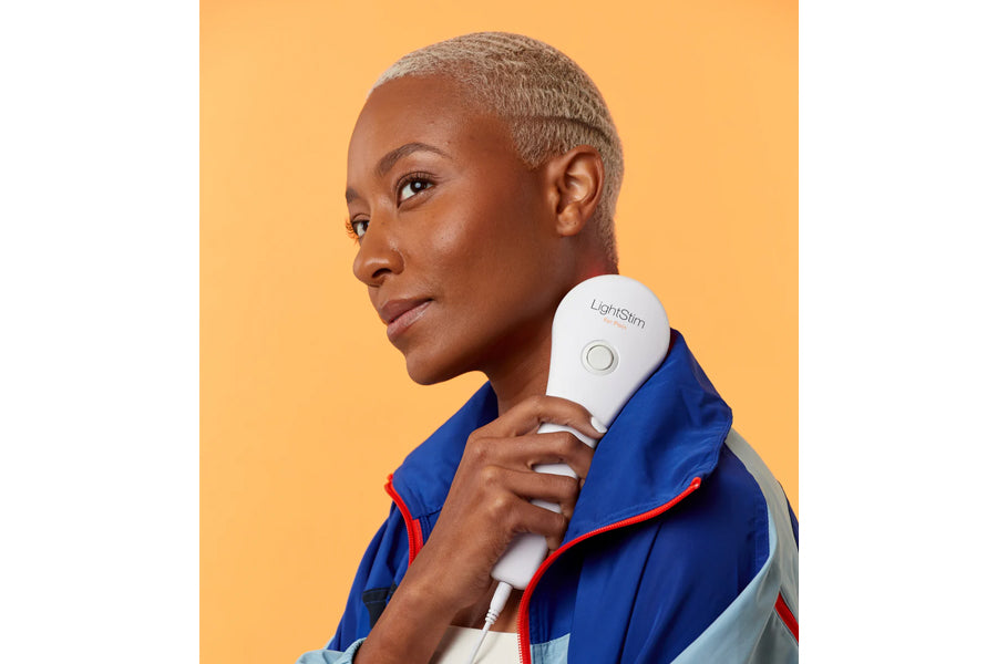 Model holding white and blue LightStim LED light therapy device against shoulder, worn with blue and red jacket on yellow bac
