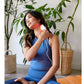 Woman holding white LED light therapy device against neck, wearing blue sleeveless top, seated with potted plants in backgrou
