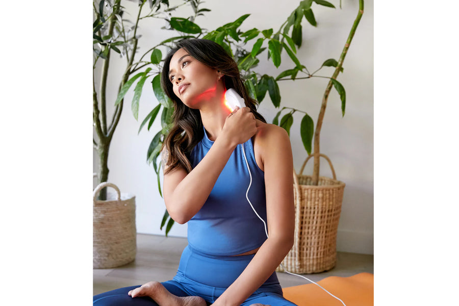 Woman holding white LED light therapy device against neck, wearing blue sleeveless top, seated with potted plants in backgrou