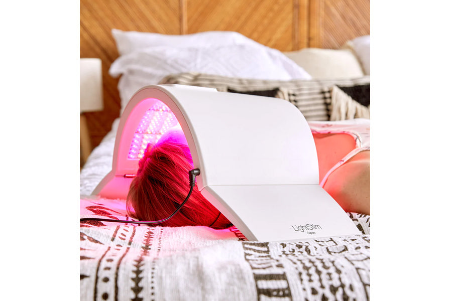 White LED light therapy lamp with pink glowing dome emitting LED light, shown on bed with model wearing treatment cap
