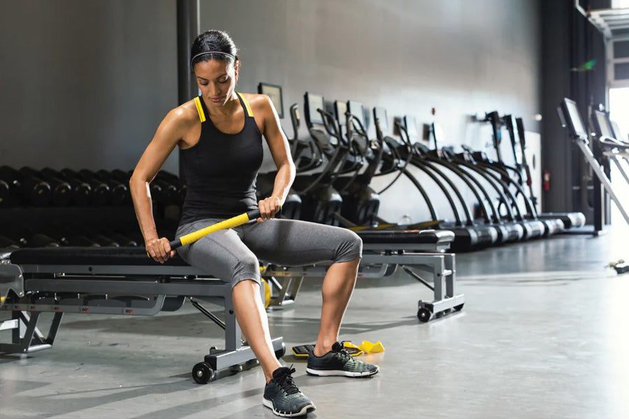 Woman using yellow and black massage bar on leg in gym setting with equipment in background