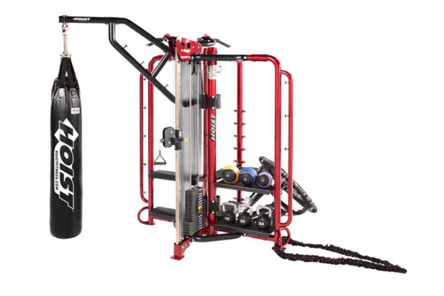 HOIST red and black functional training cage with integrated workout station, hanging heavy bag, and adjustable dumbbells on 