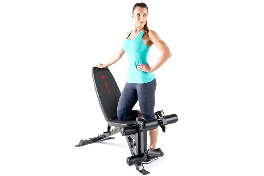 Woman wearing turquoise tank top and navy capri leggings posing next to black Marcy Deluxe Utility Weight Bench with red logo