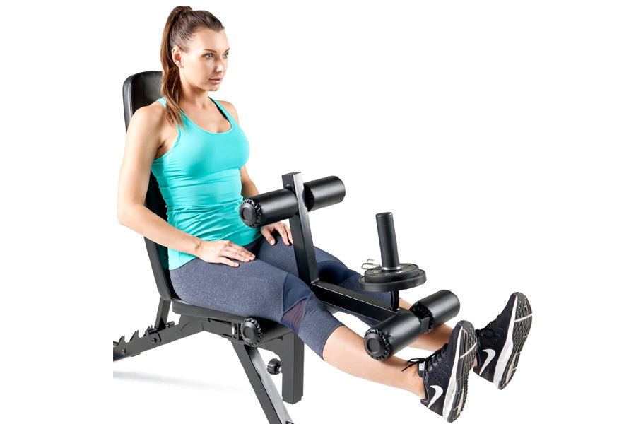 Woman in turquoise tank top and navy leggings seated on black leg extension weight bench with foot pedals, holding dumbbell, 