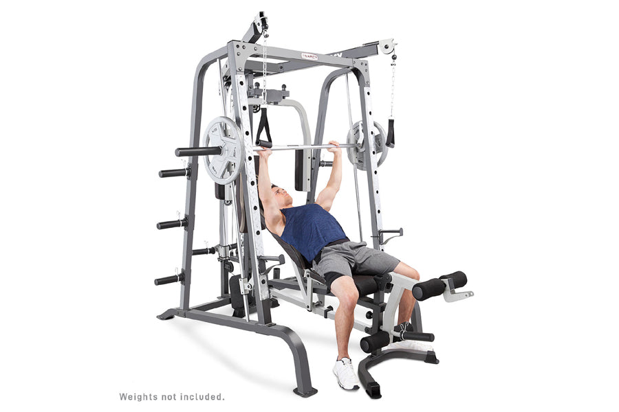 Man in navy shirt and gray shorts using white and gray Smith machine cage system with barbell on white background