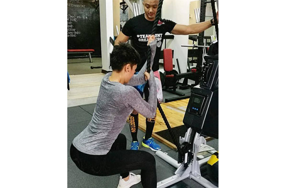 Woman using gray long-sleeve top exercising on Marpo VMX Rope Trainer in gym setting with instructor assisting