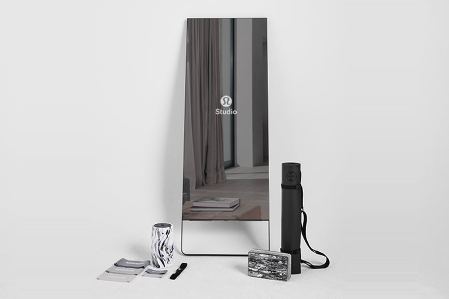 Lululemon Studio Mirror Plus with black frame standing upright, displaying studio reflection, surrounded by accessories inclu