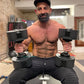 Man holding black adjustable dumbbells in home gym setting, wearing black cap and shorts