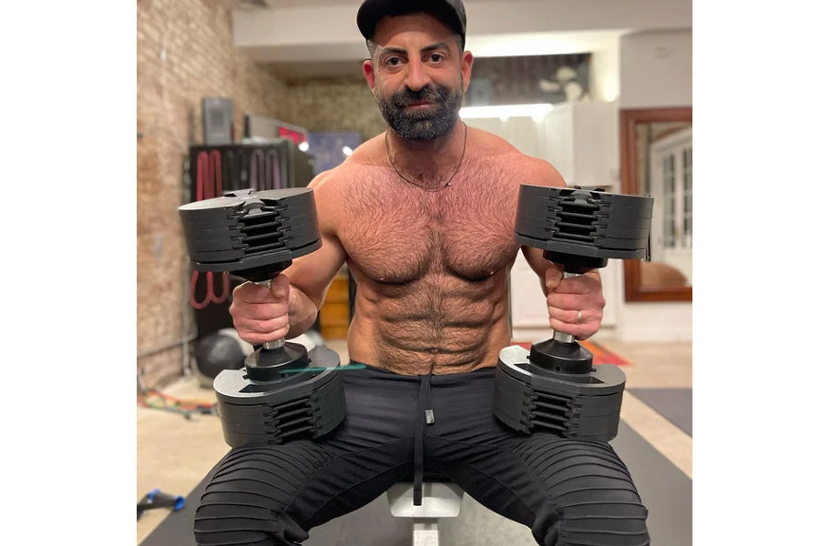 Man holding black adjustable dumbbells in home gym setting, wearing black cap and shorts