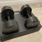 Pair of matte black adjustable dumbbells with numbered weight selector dials on a black storage tray, on gray background
