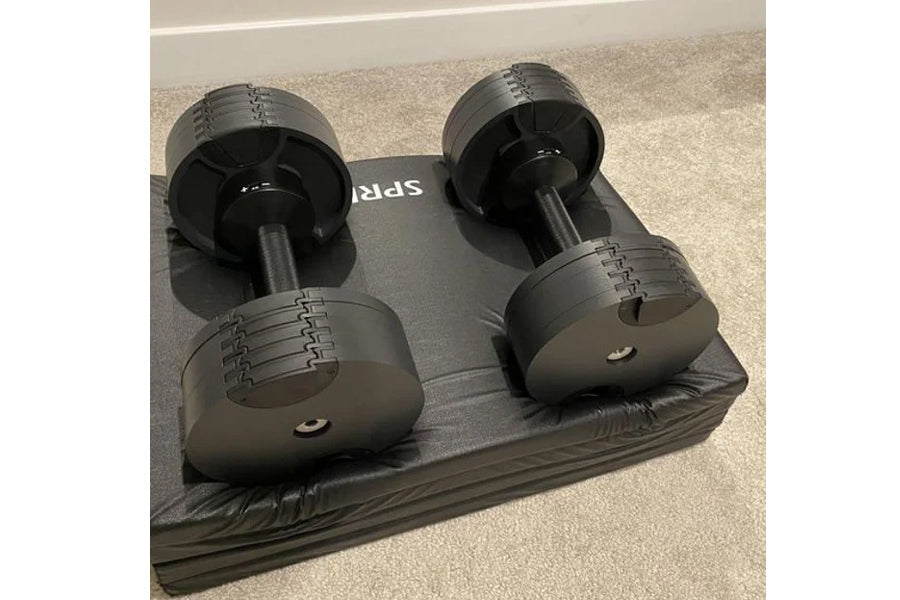 Pair of matte black adjustable dumbbells with numbered weight selector dials on a black storage tray, on gray background