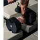 Black adjustable dumbbell with neon yellow accents held by model wearing black mesh sneaker with neon trim on concrete surfac