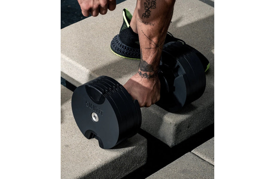 Black adjustable dumbbell with neon yellow accents held by model wearing black mesh sneaker with neon trim on concrete surfac