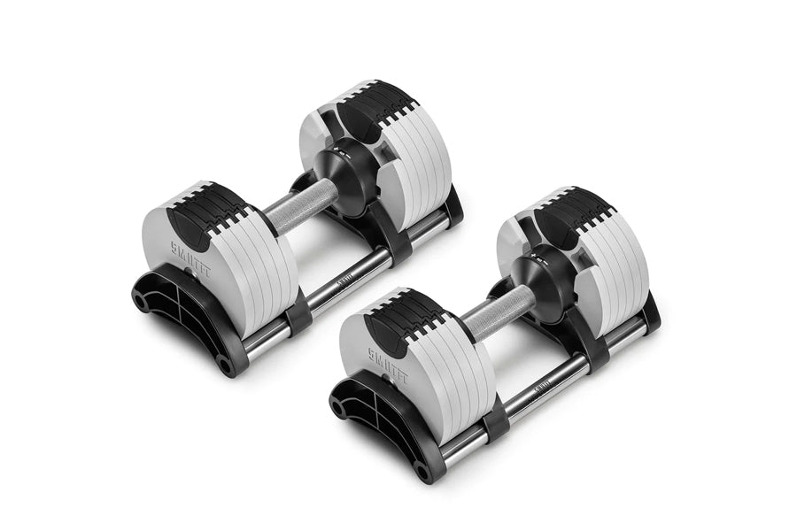 Pair of NÜOBELL adjustable dumbbells with white and black weight plates, chrome handles, and black base stands on white backg