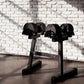 Black adjustable dumbbell stand with double legs holding multiple black dumbbells against white brick wall