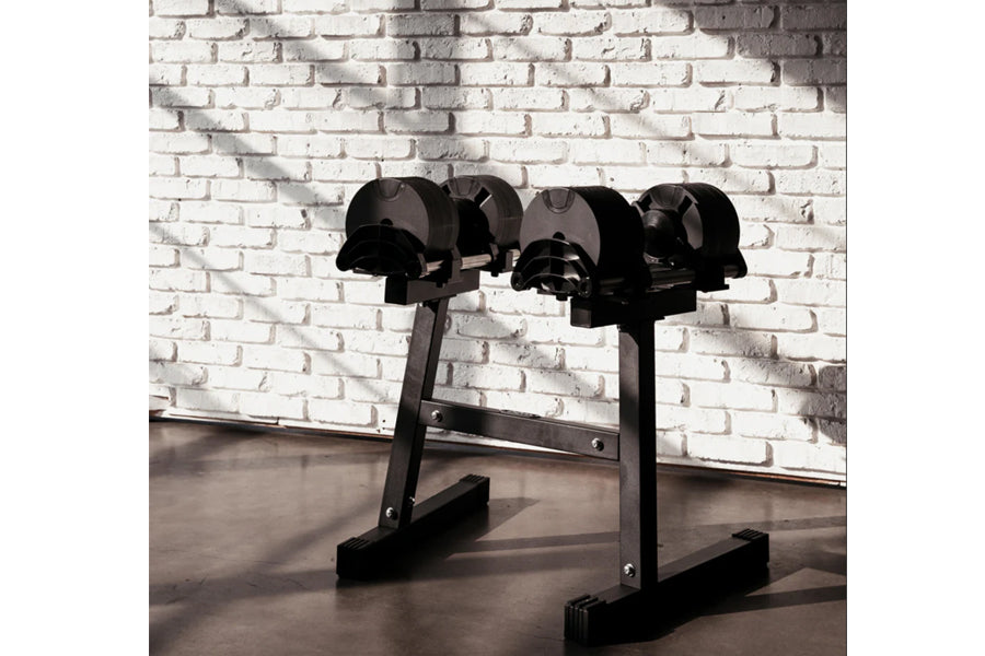 Black adjustable dumbbell stand with double legs holding multiple black dumbbells against white brick wall