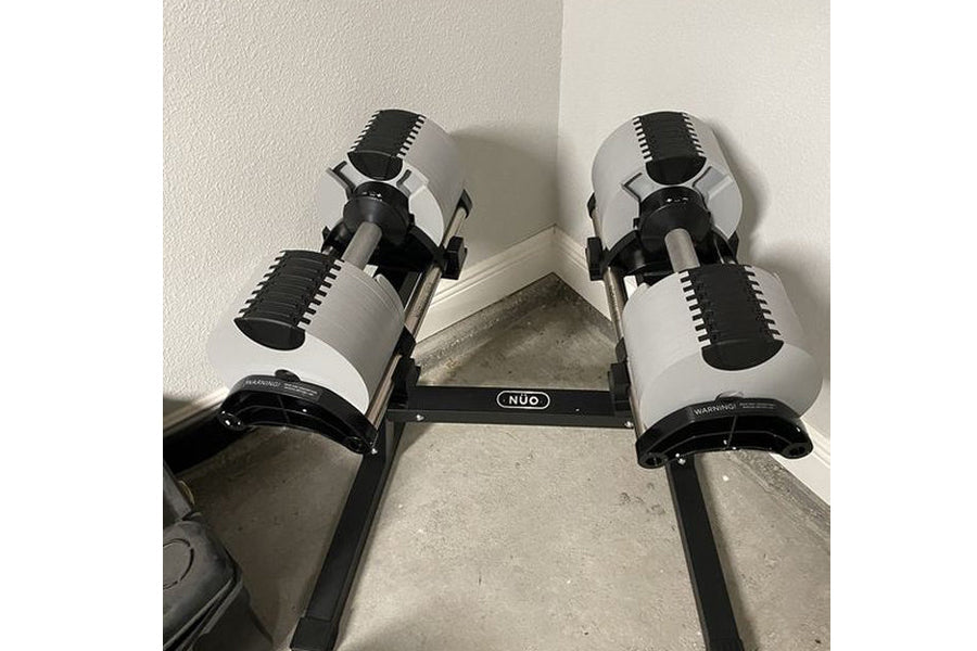 Black and white adjustable dumbbell stand with two dumbbells, NÜOBELL branding visible on horizontal bar, on concrete floor