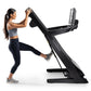 Woman wearing gray sports bra and black leggings running on black NordicTrack treadmill with digital display against white ba