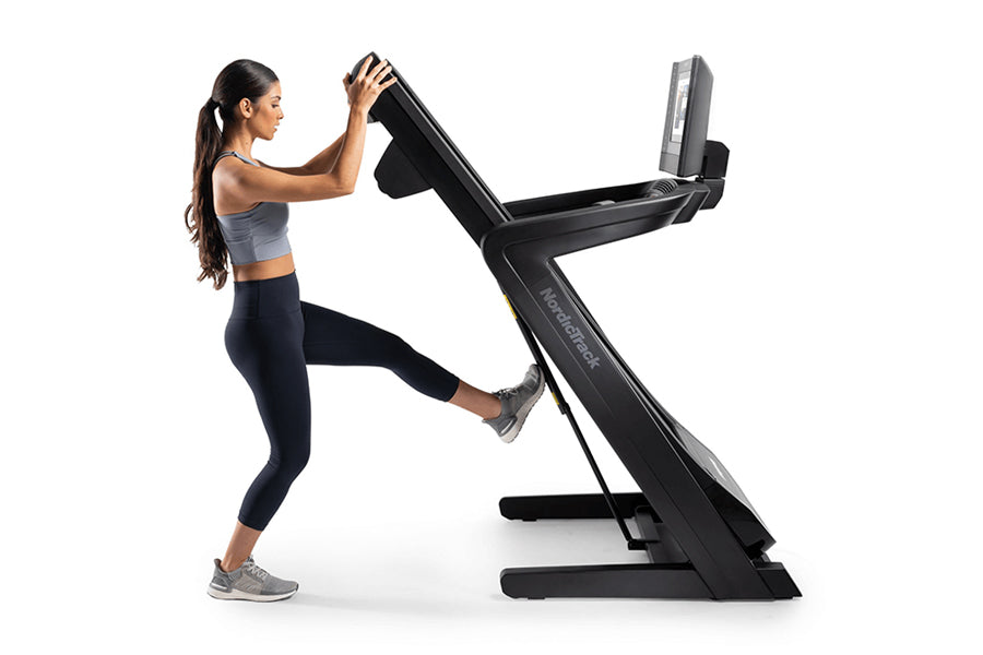 Woman wearing gray sports bra and black leggings running on black NordicTrack treadmill with digital display against white ba