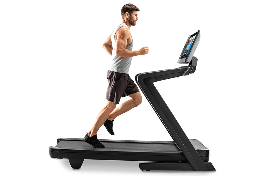 Man running on black NordicTrack treadmill with digital display, wearing gray tank top and black shorts
