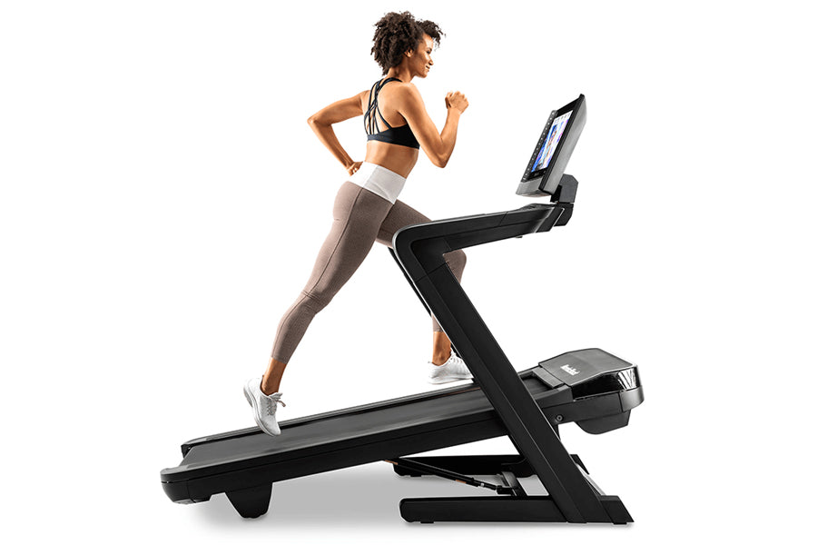 Woman running on black NordicTrack treadmill with digital display, wearing black sports bra and taupe leggings