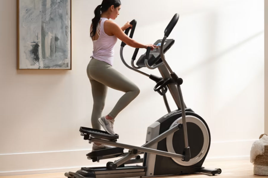 Woman wearing lavender tank top and olive leggings using black and gray elliptical machine in bright home gym setting