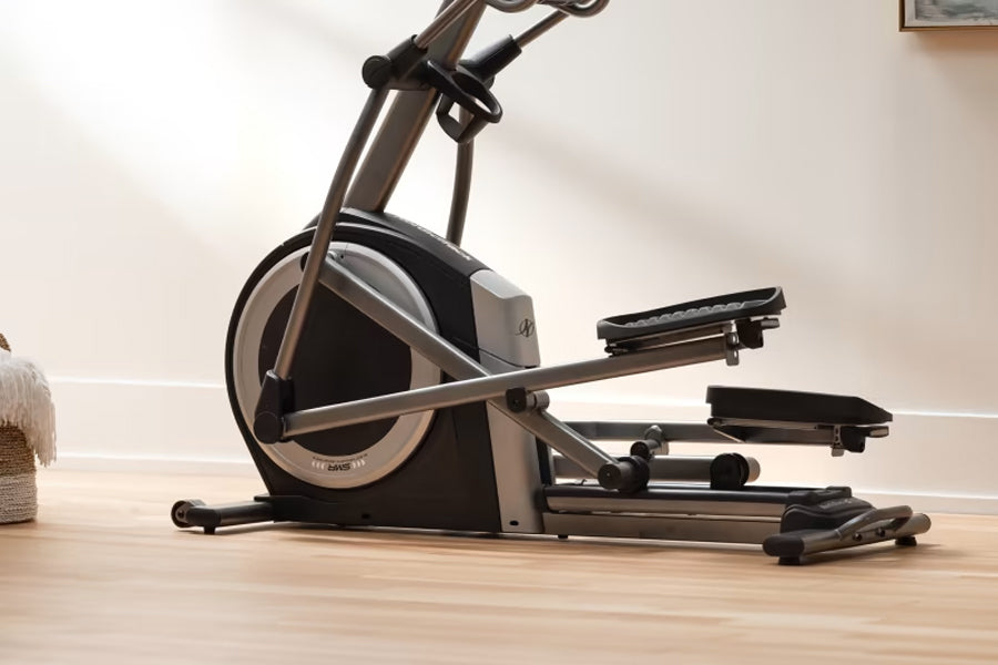 Black and gray commercial elliptical machine with large wheel and dual footpeds on wooden floor against white wall